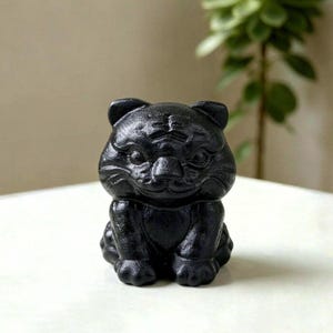 Könnte beinhalten: Eine kleine, massive schwarze Gusseisenfigur in Form eines sitzenden Tigers. Der Tiger hat detaillierte Gesichtszüge, darunter spitze Ohren, definierte Augen, eine Nase und Schnurrhaare, mit einer strukturierten Oberfläche am ganzen Körper.