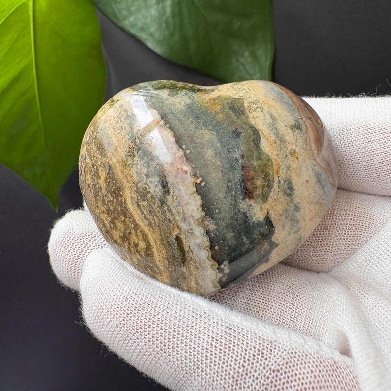 May include: A heart-shaped polished stone featuring a complex pattern of earthy tones including dark green, tan, beige, and cream. The surface displays natural banding and mineral inclusions with a smooth, glossy finish that reflects light.