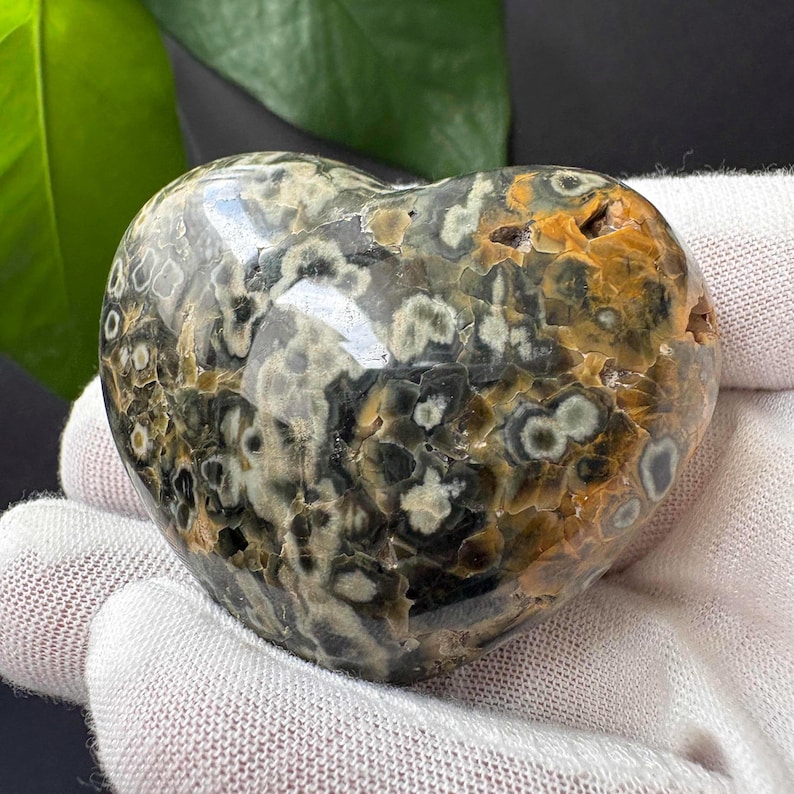 May include: A polished heart-shaped stone featuring a complex pattern of dark grey, black, and mustard yellow circular inclusions. The surface is smooth and glossy, displaying a variety of organic, flower-like spots throughout the entire object.