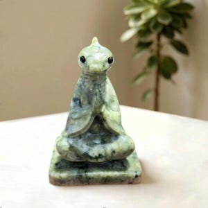 Può includere: Una piccola statuetta in pietra intagliata a mano raffigurante una creatura con una superficie liscia, verde pallido e screziata di scuro. La figura è raffigurata in una posa seduta e meditativa con le mani giunte davanti al petto e due piccoli occhi neri.