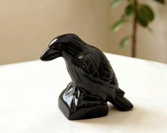 May include: A polished black stone carving of a raven perched on a base. The sculpture features smooth, reflective surfaces with incised lines detailing the bird's feathers, wings, and beak, capturing the form of the bird in a resting position.