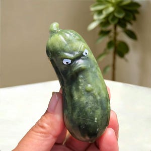 May include: A dark green, smooth, stone-like sculpture shaped like a cucumber with a bumpy texture. The object features a carved, angry facial expression with two white eyes and black pupils, giving it a grumpy appearance with a furrowed brow.