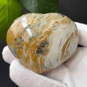 May include: A polished heart-shaped stone featuring a swirling pattern of mustard yellow, slate grey, and creamy white colours. The surface is smooth and glossy, displaying intricate natural banding and mineral inclusions throughout the entire object.