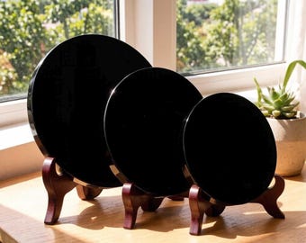May include: Three circular, solid black display plates of varying sizes, each resting on a dark reddish-brown wooden stand. The plates have a smooth, reflective surface and are arranged in a row from largest to smallest on a light wood surface.