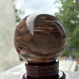 Puede incluir: Una piedra pulida y esférica con un patrón arremolinado de tonos marrones y beige. La esfera se exhibe sobre un soporte de madera oscura, mostrando su belleza natural. La superficie de la piedra refleja la luz.