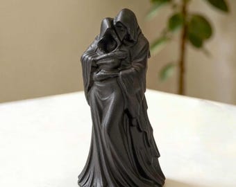 May include: A matte black figurine depicting two hooded figures in a close embrace. The figures are draped in long, flowing robes with visible folds and textures that extend to the base. The sculpture features a unified, dark aesthetic.