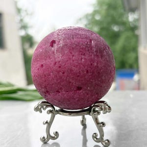 May include: A round, textured, deep magenta sphere, possibly a gemstone, sits atop an ornate silver metal stand. The sphere has small, irregular surface imperfections. The stand features a decorative, scrolled design.