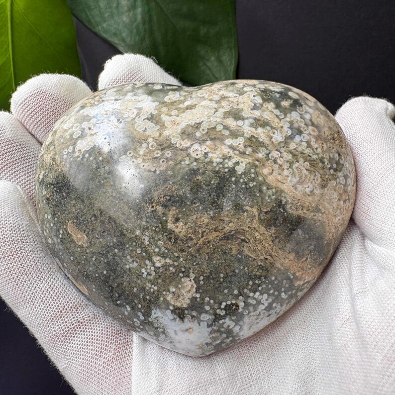 May include: A polished heart-shaped stone featuring a mottled pattern of dark forest green, muted grey, and creamy beige tones. The surface displays intricate circular orbicular inclusions and swirling mineral veins throughout the smooth, rounded form.