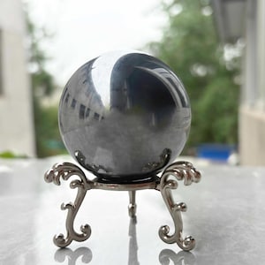 Può includere: Una sfera lucida, grigio scuro, poggia su un supporto in metallo argentato ornato con tre gambe curve. La sfera riflette l'ambiente circostante, creando un effetto a specchio. Il supporto presenta un design decorativo a volute.