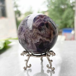 May include: A polished amethyst sphere, displaying shades of purple and white, rests on an ornate silver-colored metal stand. The sphere's surface reflects light, showcasing its natural patterns. The stand has a decorative, three-legged design.