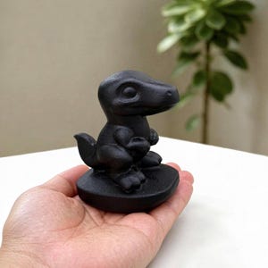 May include: A small, matte black figurine of a dinosaur with a large head, prominent eyes, and short arms, standing on a rounded base. The creature is depicted in a seated position with its tail curled behind it, featuring a smooth, solid surface.