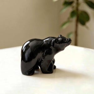 Könnte beinhalten: Eine kleine, massive schwarze Bärenfigur mit einer glatten, glänzenden Oberfläche. Der Bär ist in stehender Position dargestellt, nach rechts gewandt mit leicht nach oben geneigtem Kopf, was detaillierte Schnitzereien am Körper zeigt.