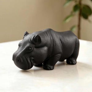 Könnte beinhalten: Eine mattschwarze Keramikfigur eines Flusspferdes, das in Seitenansicht steht. Die Skulptur weist eine texturierte Oberfläche, abgerundete Ohren, eine breite Schnauze und vier kräftige Beine auf und zeigt das Tier in einer festen, ruhigen Haltung.