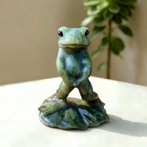 May include: A small, hand-carved green stone figurine of a frog standing upright on a lily pad. The sculpture features a smooth, polished surface with natural dark green and olive marbling throughout, showcasing a detailed frog form.