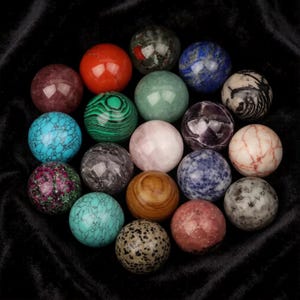 May include: A selection of gemstone spheres in a variety of colours and patterns. The spheres include red, green, blue, purple, and brown hues, with textures from smooth to marbled. The arrangement is displayed on a dark, velvety surface.
