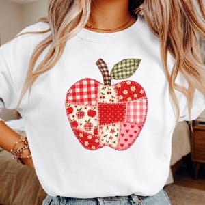 May include: A white crewneck sweatshirt featuring a large apple graphic composed of various red, white, and green fabric-style patches. The apple design includes patterns of gingham, polka dots, small flowers, and hearts, with a brown checkered stem.