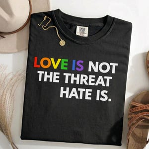 May include: Black t-shirt with the text "LOVE IS NOT THE THREAT HATE IS." The word "LOVE" is in rainbow colors. A gold necklace and a beige hat are visible in the background.