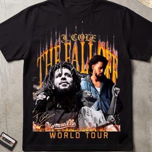 May include: A black short-sleeve t-shirt featuring a graphic print of J. Cole. The design includes the text J. COLE, THE FALL OFF, and WORLD TOUR in orange and yellow, with multiple images of the artist and a signature on the front.