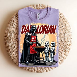 May include: A lilac t-shirt with a graphic of Darth Vader grilling with Stormtroopers. The text "Darthlorian" is above the image, and Darth Vader wears a red apron that says "#1 Dad".