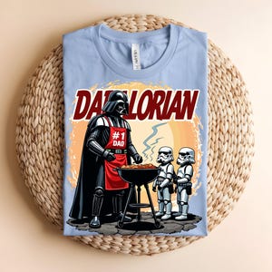 May include: Pale blue t-shirt with a graphic of Darth Vader wearing an apron that says "#1 Dad" grilling with two Stormtroopers. The text "Darlorian" is above the image. The shirt is on a woven mat.