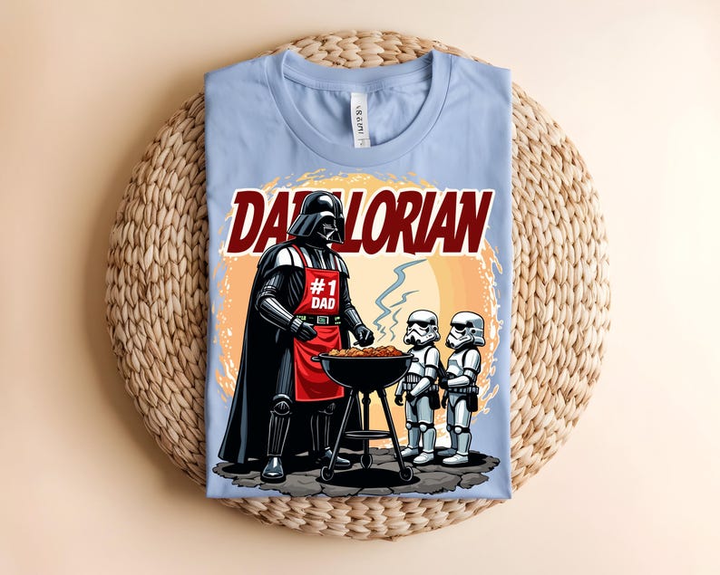 May include: Pale blue t-shirt with a graphic of Darth Vader wearing an apron that says "#1 Dad" grilling with two Stormtroopers. The text "Darlorian" is above the image. The shirt is on a woven mat.
