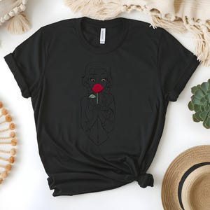 May include: Black t-shirt featuring a line drawing of a person holding a red rose. The graphic is centered on the shirt. The shirt has a crew neck and short sleeves. The shirt is made of a soft material.