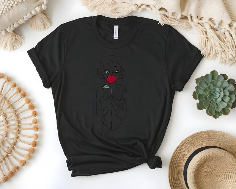 May include: Black t-shirt featuring a line drawing of a person holding a red rose. The graphic is centered on the shirt. The shirt has a crew neck and short sleeves. The shirt is made of a soft material.