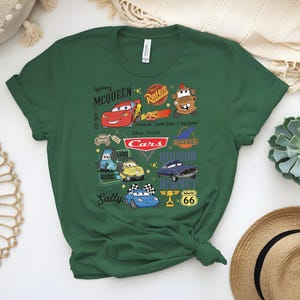 May include: Green t-shirt featuring characters from the Disney Pixar Cars movie. The design includes Lightning McQueen, Mater, and other characters, along with the Cars logo and Route 66 signage. The shirt is made of a soft material.