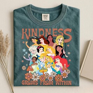 May include: Teal Comfort Colors t-shirt featuring a graphic of various cartoon princesses. The word "KINDNESS" is printed above the princesses, and "GROWS FROM WITHIN" is printed below. The design includes flowers and mushrooms.