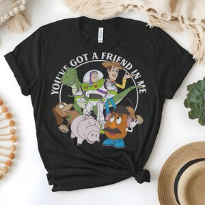 May include: Black crew neck t-shirt with a circular graphic featuring the phrase "YOU'VE GOT A FRIEND IN ME" and cartoon characters. The characters include a green dinosaur, Buzz Lightyear, Woody, a pig, and Mr. Potato Head.