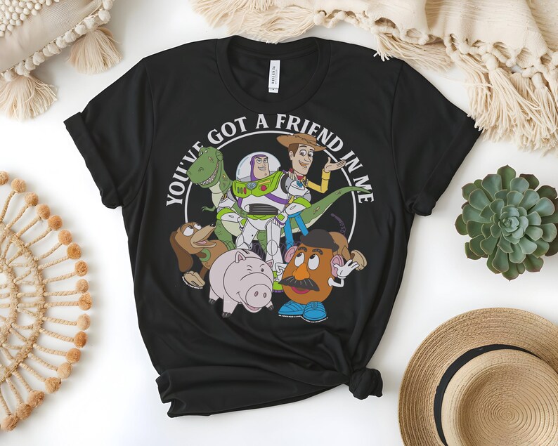 May include: Black crew neck t-shirt with a circular graphic featuring the phrase "YOU'VE GOT A FRIEND IN ME" and cartoon characters. The characters include a green dinosaur, Buzz Lightyear, Woody, a pig, and Mr. Potato Head.