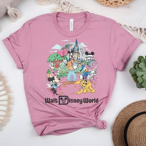 Puede incluir: Una camiseta rosa polvorienta con un gr&aacute;fico colorido de personajes y lugares emblem&aacute;ticos de Disney. El dise&ntilde;o incluye Mickey y Minnie Mouse, Goofy, Donald Duck y el texto "Walt Disney World". La camiseta es de material suave.
