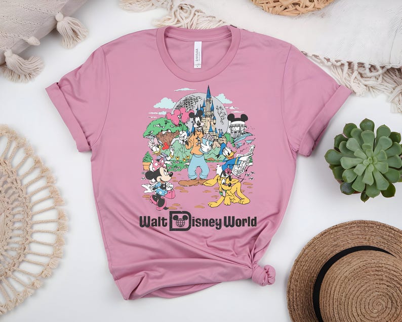 Puede incluir: Una camiseta rosa polvorienta con un gr&aacute;fico colorido de personajes y lugares emblem&aacute;ticos de Disney. El dise&ntilde;o incluye Mickey y Minnie Mouse, Goofy, Donald Duck y el texto "Walt Disney World". La camiseta es de material suave.