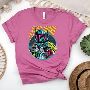 May include: A pink t-shirt with a graphic of a Star Wars character in a helmet, holding a weapon. The character is set in a blue circle with the words "STAR WARS" in yellow and white. The shirt is made of soft material.