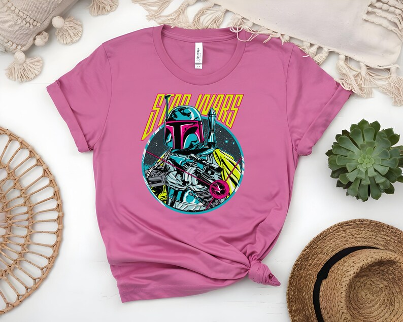 May include: A pink t-shirt with a graphic of a Star Wars character in a helmet, holding a weapon. The character is set in a blue circle with the words "STAR WARS" in yellow and white. The shirt is made of soft material.
