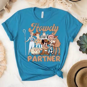 May include: A turquoise t-shirt featuring a graphic of cartoon characters with the text "Howdy Partner" in a retro font. The design includes characters like Woody, Buzz Lightyear, and Bo Peep. The shirt is knotted at the bottom.