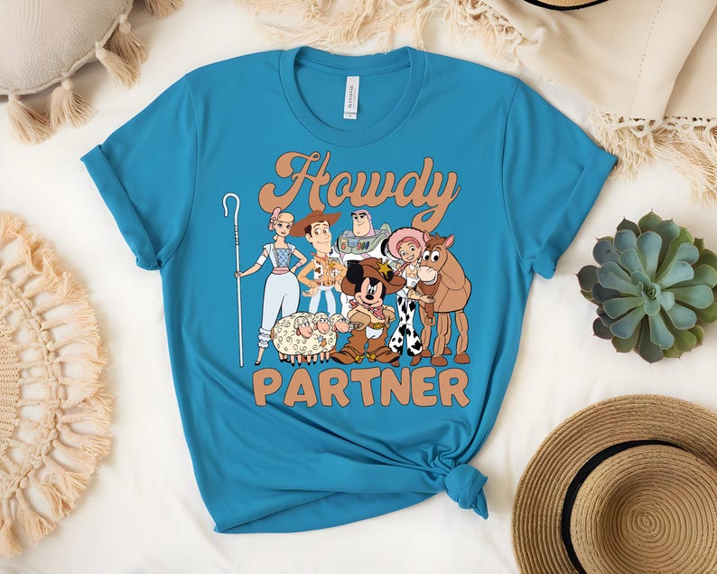 May include: A turquoise t-shirt featuring a graphic of cartoon characters with the text "Howdy Partner" in a retro font. The design includes characters like Woody, Buzz Lightyear, and Bo Peep. The shirt is knotted at the bottom.