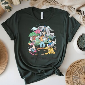 Puede incluir: Camiseta verde oscuro con un gr&aacute;fico colorido de personajes de Disney, incluyendo Mickey Mouse, Donald Duck y Goofy, frente a un castillo y el texto "Walt Disney World". La camiseta est&aacute; hecha de material suave.