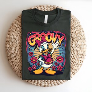 May include: Dark green t-shirt featuring a cartoon Daisy Duck design with the word "GROOVY" in a retro font. Daisy is wearing a pink bow and vest, surrounded by flowers and a burst of stars.