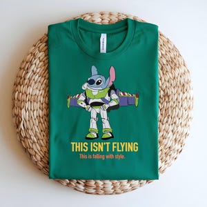 May include: Green t-shirt featuring a cartoon character in a Buzz Lightyear costume. The character is blue with large eyes and pink ears. The text "THIS ISN'T FLYING" and "This is falling with style" are printed in yellow and white.