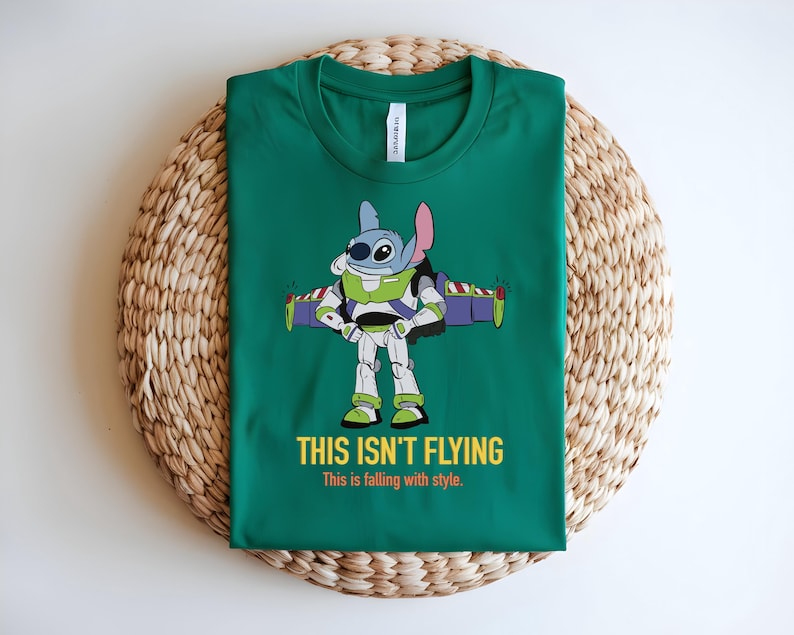May include: Green t-shirt featuring a cartoon character in a Buzz Lightyear costume. The character is blue with large eyes and pink ears. The text "THIS ISN'T FLYING" and "This is falling with style" are printed in yellow and white.