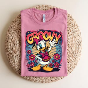 May include: A pink t-shirt featuring a cartoon Daisy Duck design with the word "GROOVY" in a retro font. Daisy is wearing a pink bow and dress, surrounded by flowers and a starburst background.