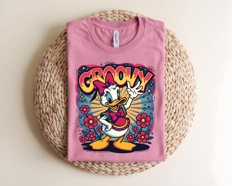 May include: A pink t-shirt featuring a cartoon Daisy Duck design with the word "GROOVY" in a retro font. Daisy is wearing a pink bow and dress, surrounded by flowers and a starburst background.