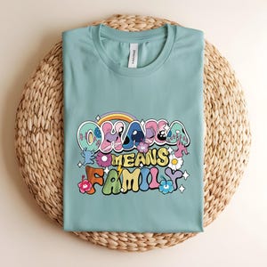 Puede incluir: Camiseta de color verde azulado claro con la frase "Ohana Means Family" en una fuente retro colorida. El dise&ntilde;o incluye un arco&iacute;ris, flores y personajes de dibujos animados. La camiseta est&aacute; doblada sobre una estera circular tejida.