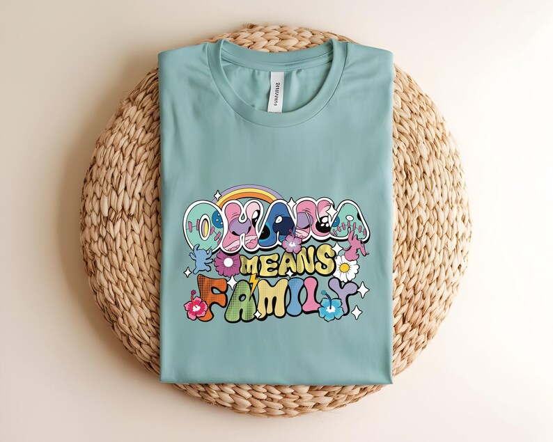 Puede incluir: Camiseta de color verde azulado claro con la frase "Ohana Means Family" en una fuente retro colorida. El dise&ntilde;o incluye un arco&iacute;ris, flores y personajes de dibujos animados. La camiseta est&aacute; doblada sobre una estera circular tejida.