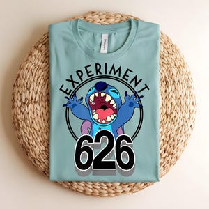 Pu&ograve; includere: Maglietta azzurra con un disegno di un personaggio dei cartoni animati. Il disegno include le parole "EXPERIMENT" e il numero "626". Il personaggio dei cartoni animati &egrave; blu con la bocca aperta e le braccia alzate, in un cerchio nero.