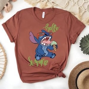K&ouml;nnte beinhalten: Rostfarbenes T-Shirt mit einem Cartoon-Charakter, Stitch, der eine Sonnenbrille tr&auml;gt und ein Eis h&auml;lt. Die Worte "hello" und "Summer" sind in gr&uuml;ner Schrift geschrieben. Das Shirt ist am unteren Rand gefaltet.