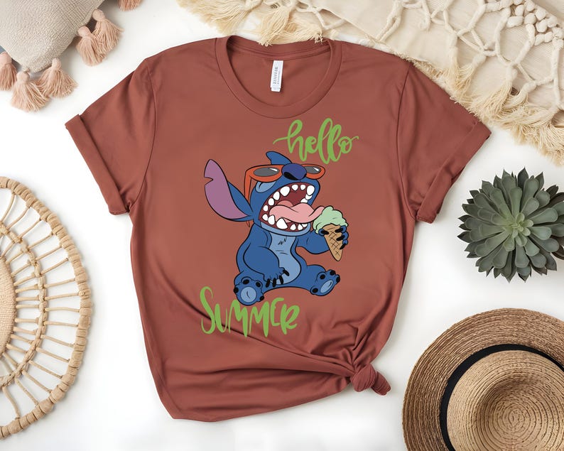 K&ouml;nnte beinhalten: Rostfarbenes T-Shirt mit einem Cartoon-Charakter, Stitch, der eine Sonnenbrille tr&auml;gt und ein Eis h&auml;lt. Die Worte "hello" und "Summer" sind in gr&uuml;ner Schrift geschrieben. Das Shirt ist am unteren Rand gefaltet.