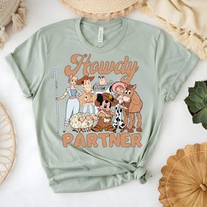 May include: Sage green t-shirt featuring a cartoon graphic of characters with the text "Howdy Partner". The graphic includes a sheep, a horse, and other characters. The shirt has a crew neck and short sleeves.