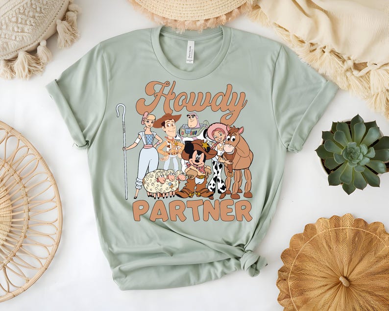 May include: Sage green t-shirt featuring a cartoon graphic of characters with the text "Howdy Partner". The graphic includes a sheep, a horse, and other characters. The shirt has a crew neck and short sleeves.